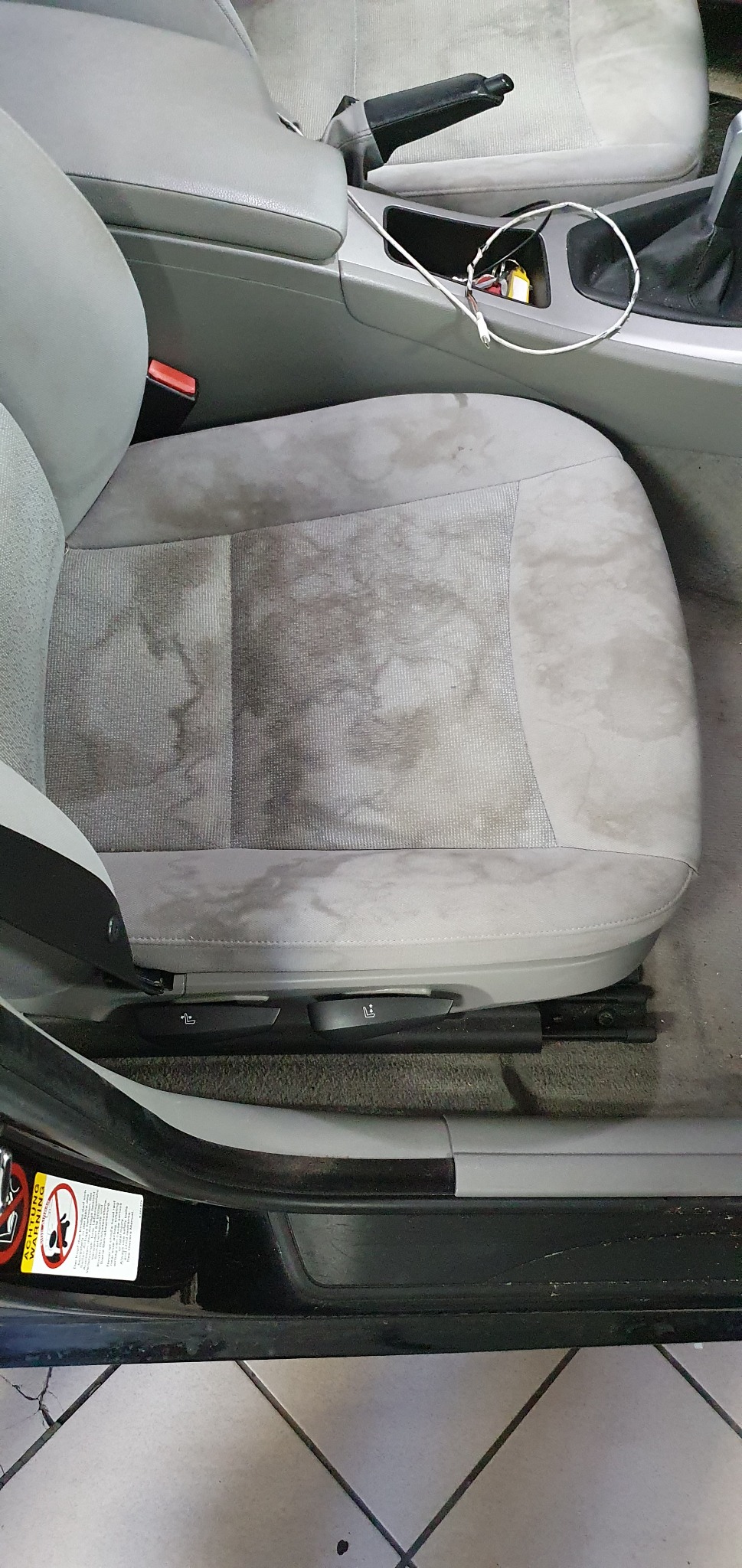 Szare, materiałowe siedzenie samochodowe z widocznymi, ciemnymi plamami i zabrudzeniami przed czyszczeniem, z boku widoczny fragment kokpitu i panelu drzwi.