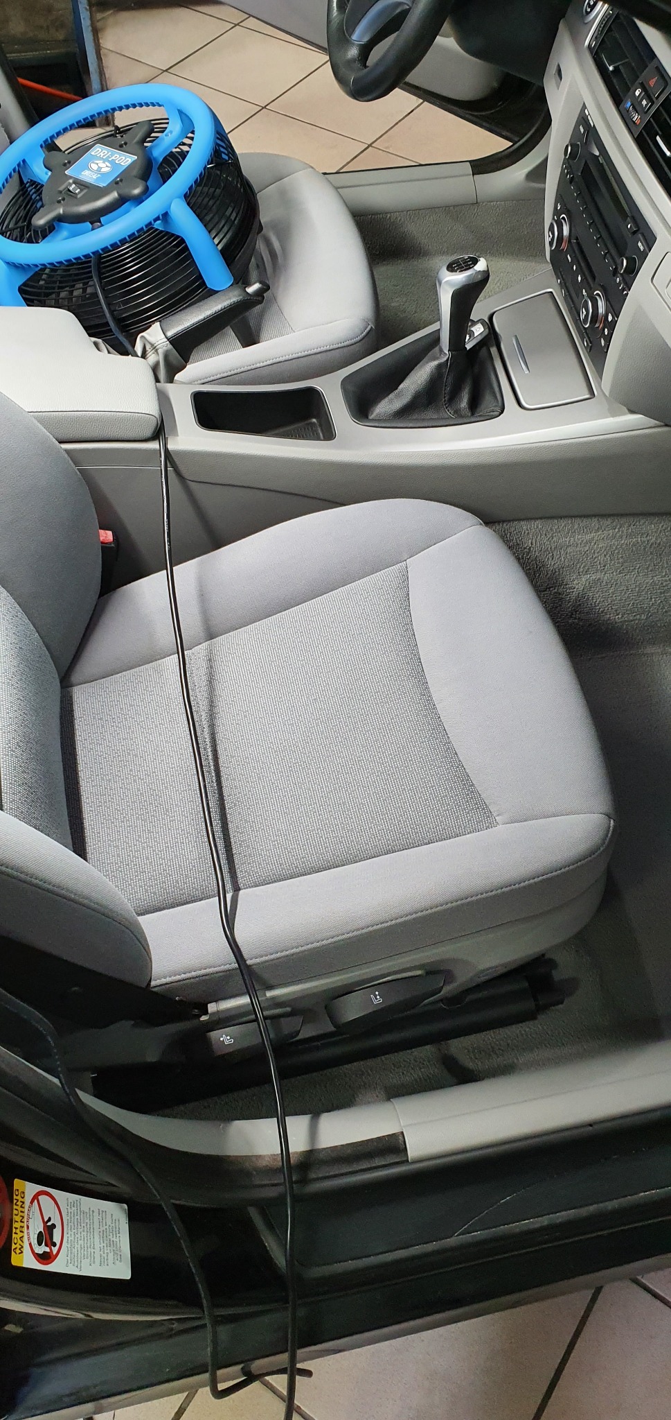 Wnętrze samochodu z szarymi fotelami poddawane suszeniu po czyszczeniu, widoczny niebieski wentylator przemysłowy na fotelu pasażera.
