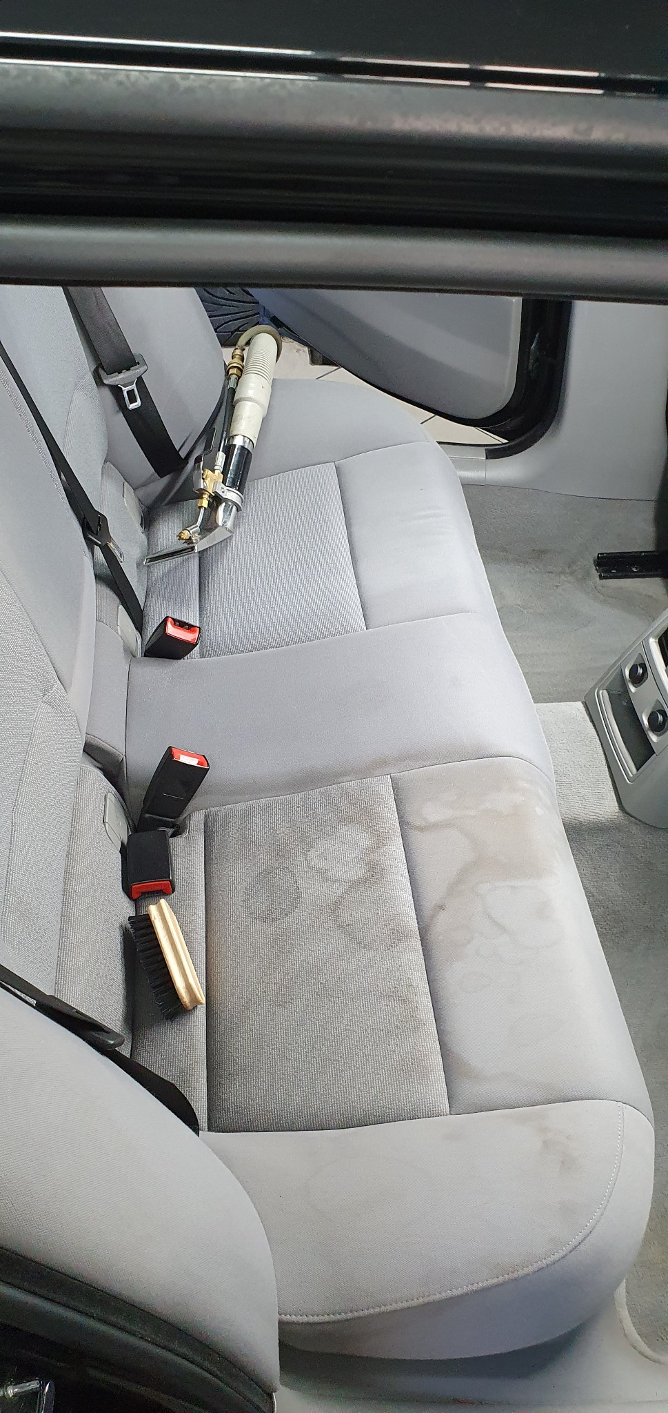 Proces czyszczenia szarej tapicerki samochodowej z widocznymi plamami, z użyciem specjalistycznego sprzętu i szczotki.