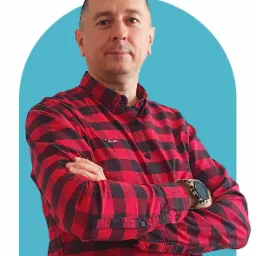 Portret mężczyzny z Rzeszowa w czerwono-czarnej koszuli w kratę, z założonymi rękami i zegarkiem na nadgarstku, na jasnoniebieskim tle.