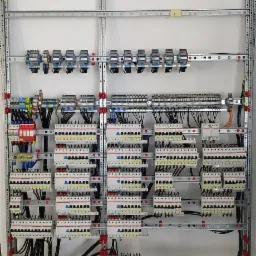 Rozbudowana rozdzielnica elektryczna zamontowana na metalowym stelażu, widoczne liczne zabezpieczenia, złączki i okablowanie, całość na tle białej ściany i surowej podłogi.