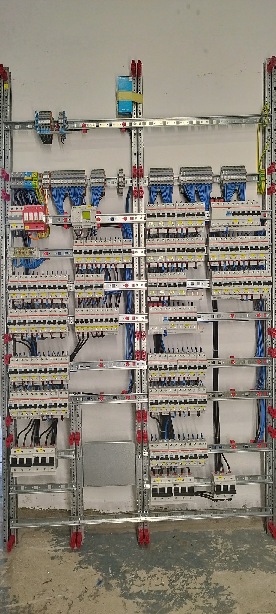 Rozbudowana instalacja elektryczna w rozdzielnicy, widoczne liczne zabezpieczenia nadprądowe, przewody w niebieskiej izolacji oraz szyny DIN zamontowane na perforowanych profilach metalowych.