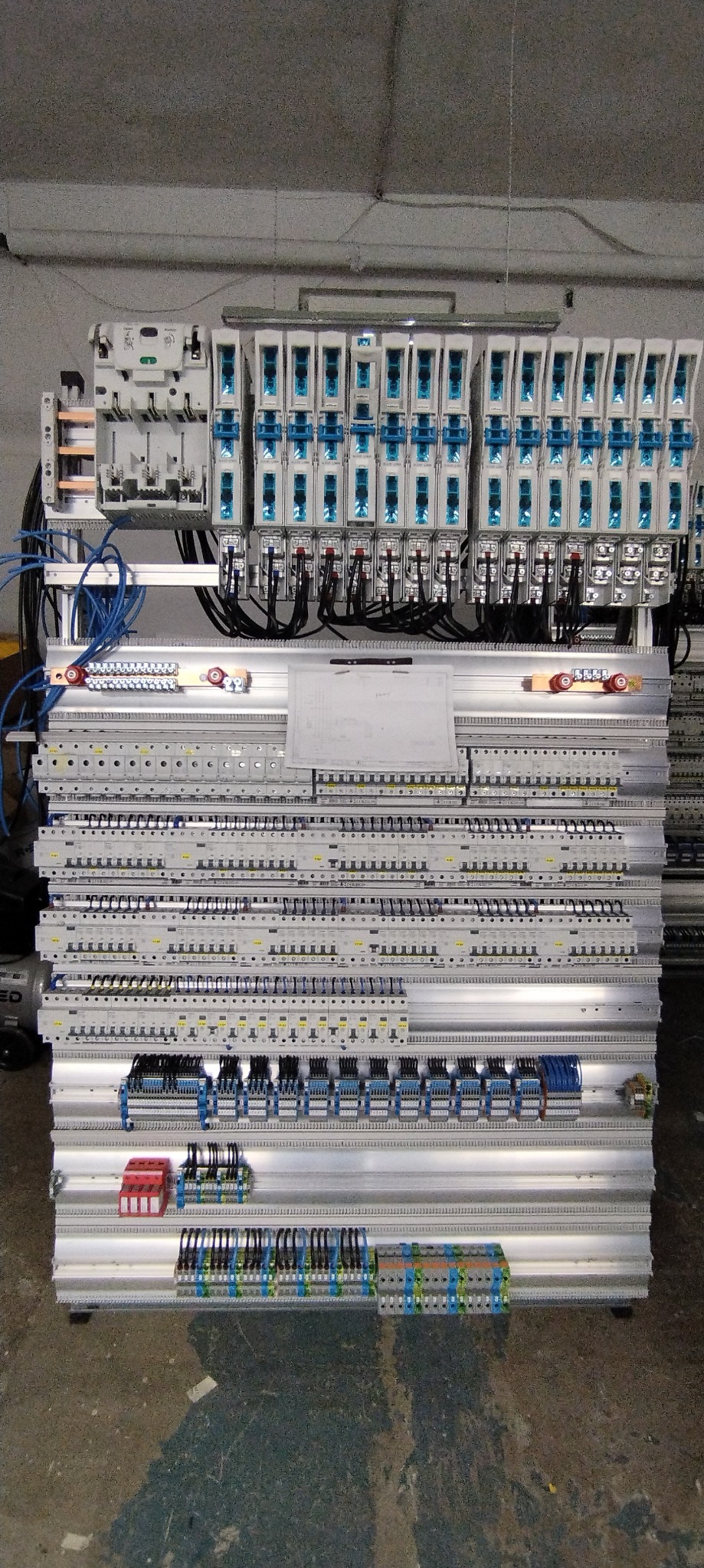 Rozbudowana, wielopoziomowa szafa sterownicza z licznymi modułami, bezpiecznikami i złączkami, okablowanie w kolorach niebieskim i czarnym, schemat umieszczony na górnym panelu, widoczne elementy...