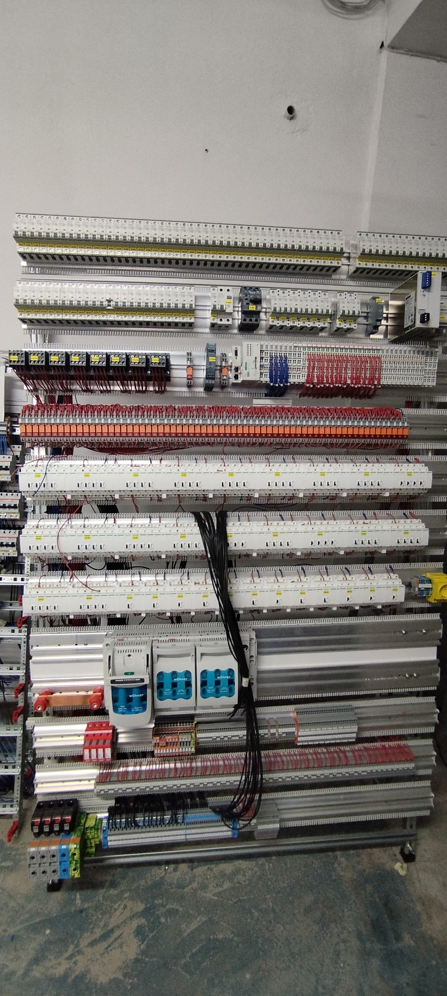 Rozbudowana szafa elektryczna z licznymi zabezpieczeniami, przewodami i modułami, zamontowana na szynach DIN, widoczne liczniki energii Wöhner.