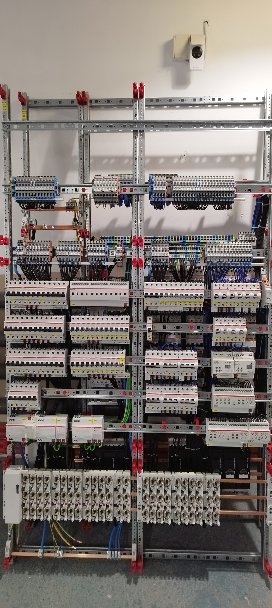 Rozbudowana, profesjonalna rozdzielnica elektryczna z licznymi zabezpieczeniami, przewodami i listwami zaciskowymi, zamontowana na metalowej konstrukcji szkieletowej w jasnym pomieszczeniu.