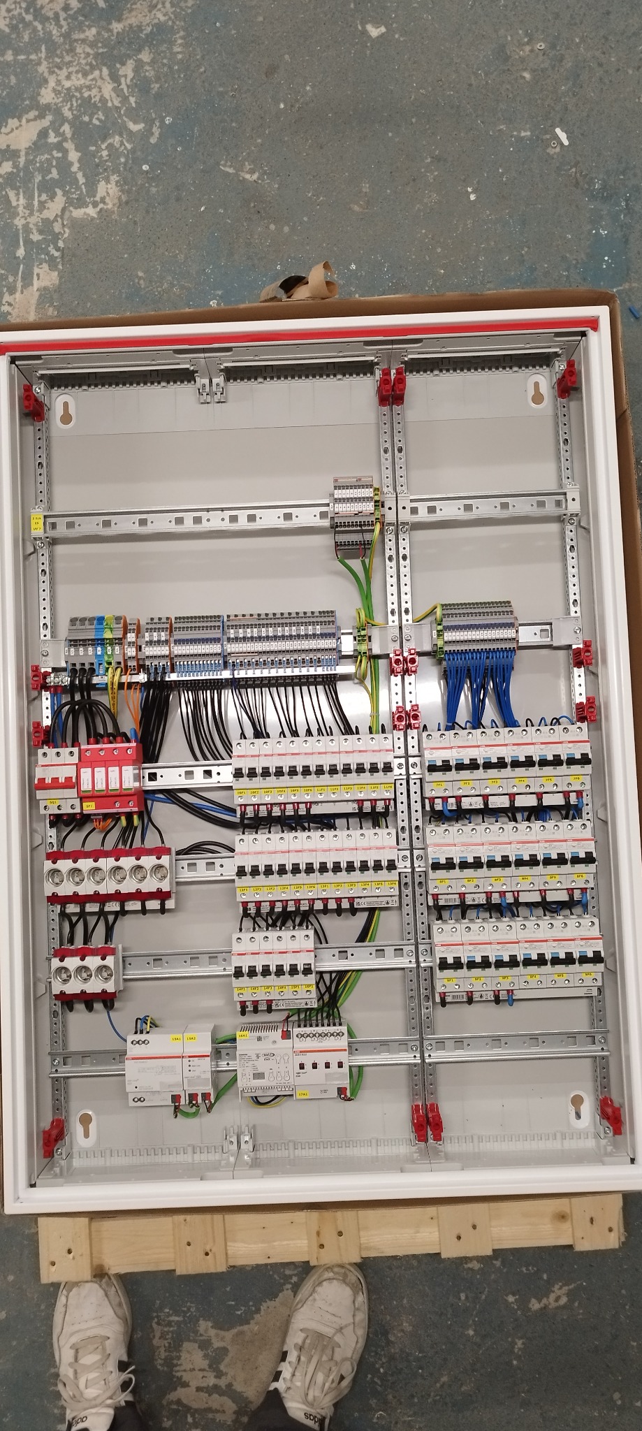 Rozdzielnica elektryczna w trakcie montażu, widoczne liczne przewody, bezpieczniki i inne elementy, całość umieszczona w białej obudowie, częściowo widoczne buty montera na tle betonowej posadzki.