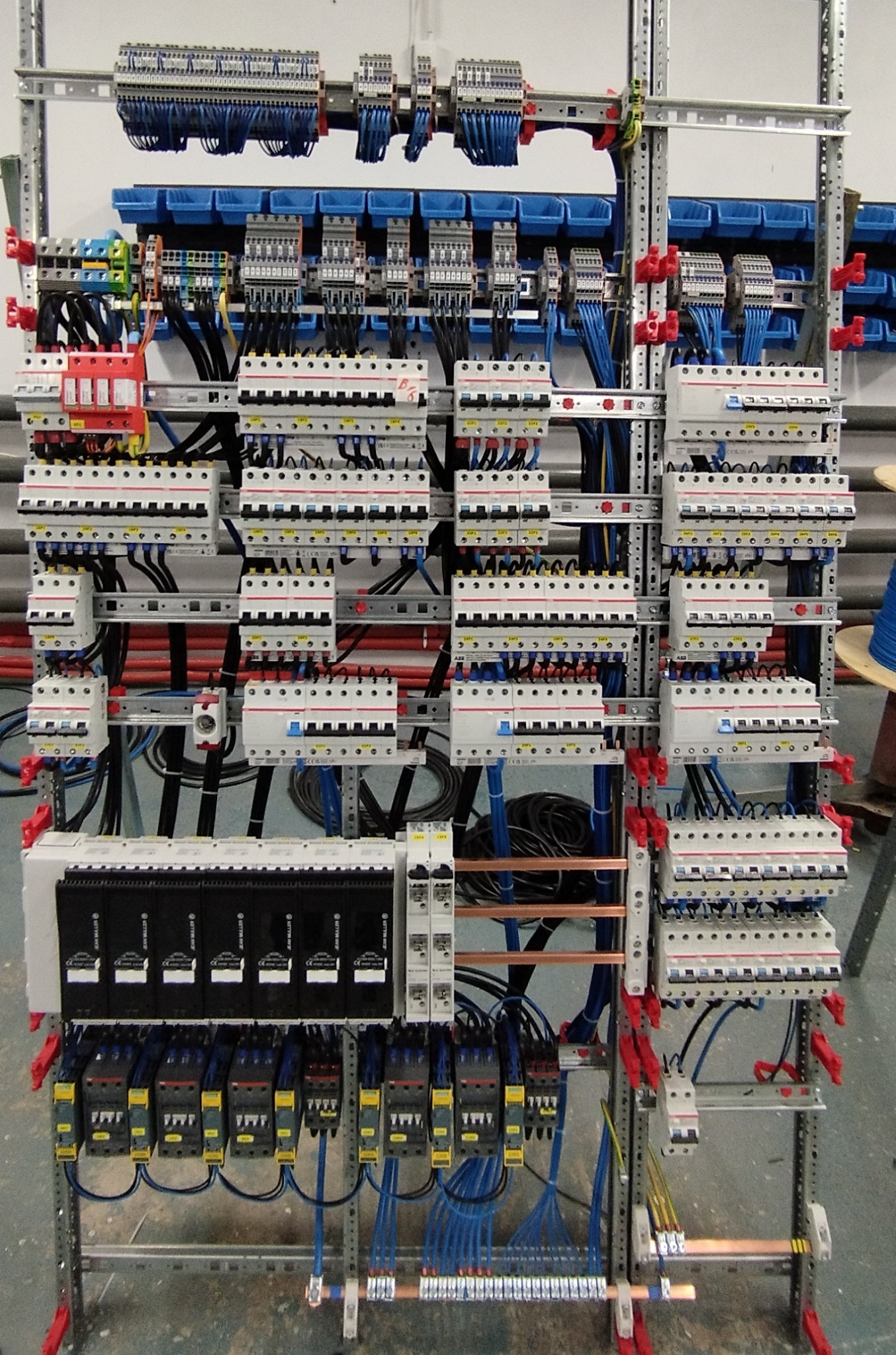 Rozbudowana, pionowa tablica elektryczna z licznymi bezpiecznikami, przekaźnikami i okablowaniem, zamontowana na metalowej konstrukcji szkieletowej z czerwonymi elementami mocującymi.