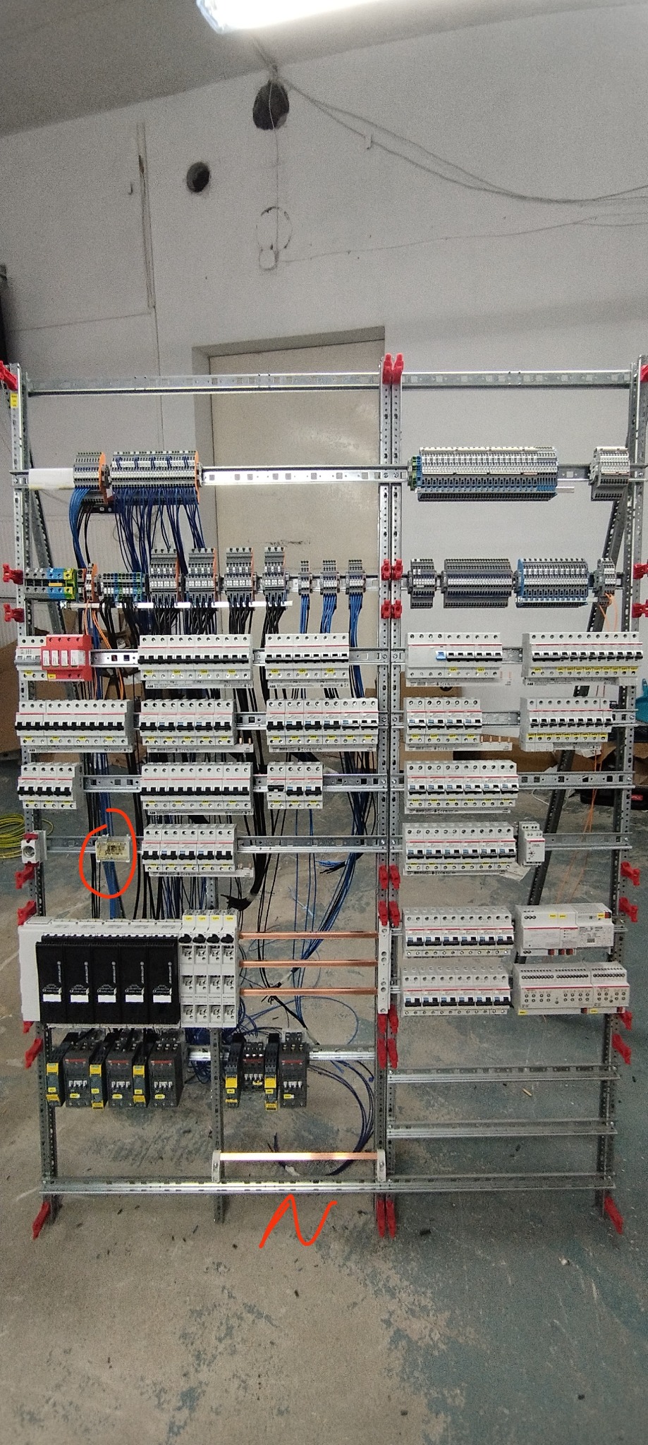 Rozdzielnica elektryczna w trakcie montażu, widoczne liczne zabezpieczenia, kable i szyny, zamontowane na metalowym stelażu, surowy beton na ścianie i podłodze w tle.