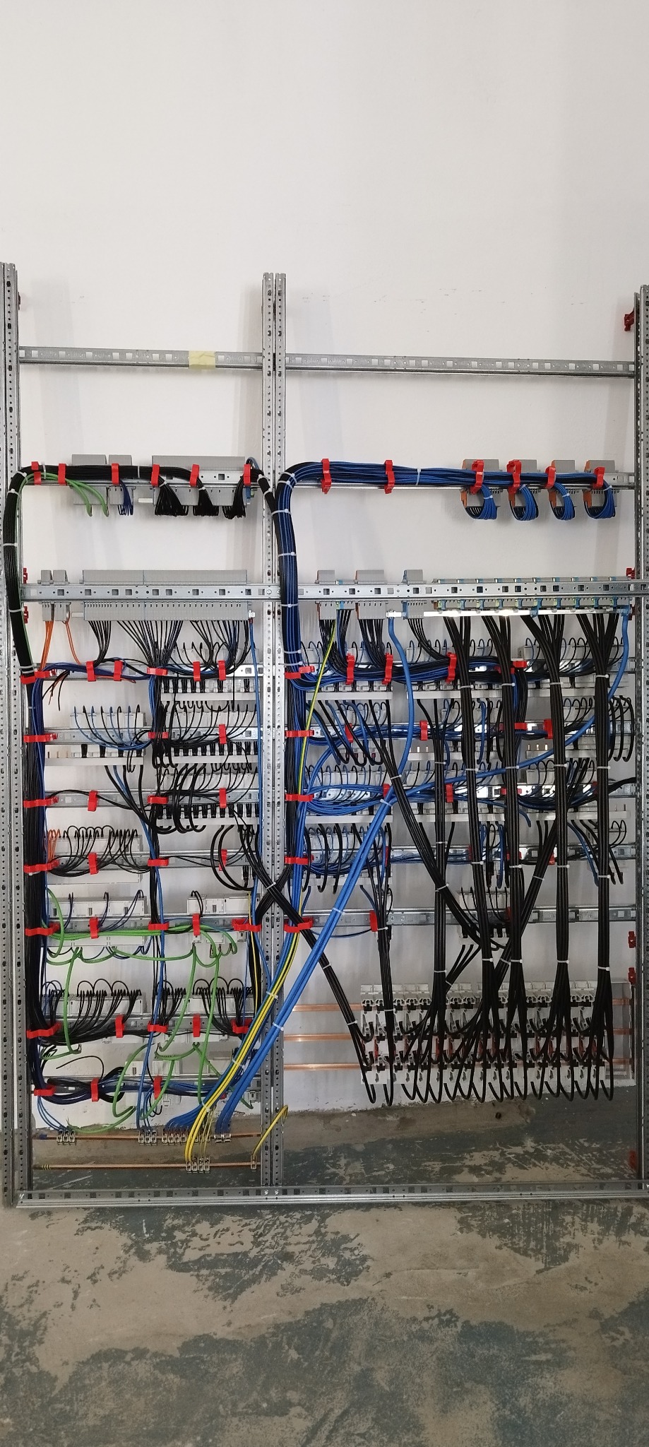 Skomplikowany system okablowania elektrycznego zamontowany na metalowej konstrukcji, z widocznymi kablami w różnych kolorach zabezpieczonych czerwonymi uchwytami, na tle białej ściany i surowej...