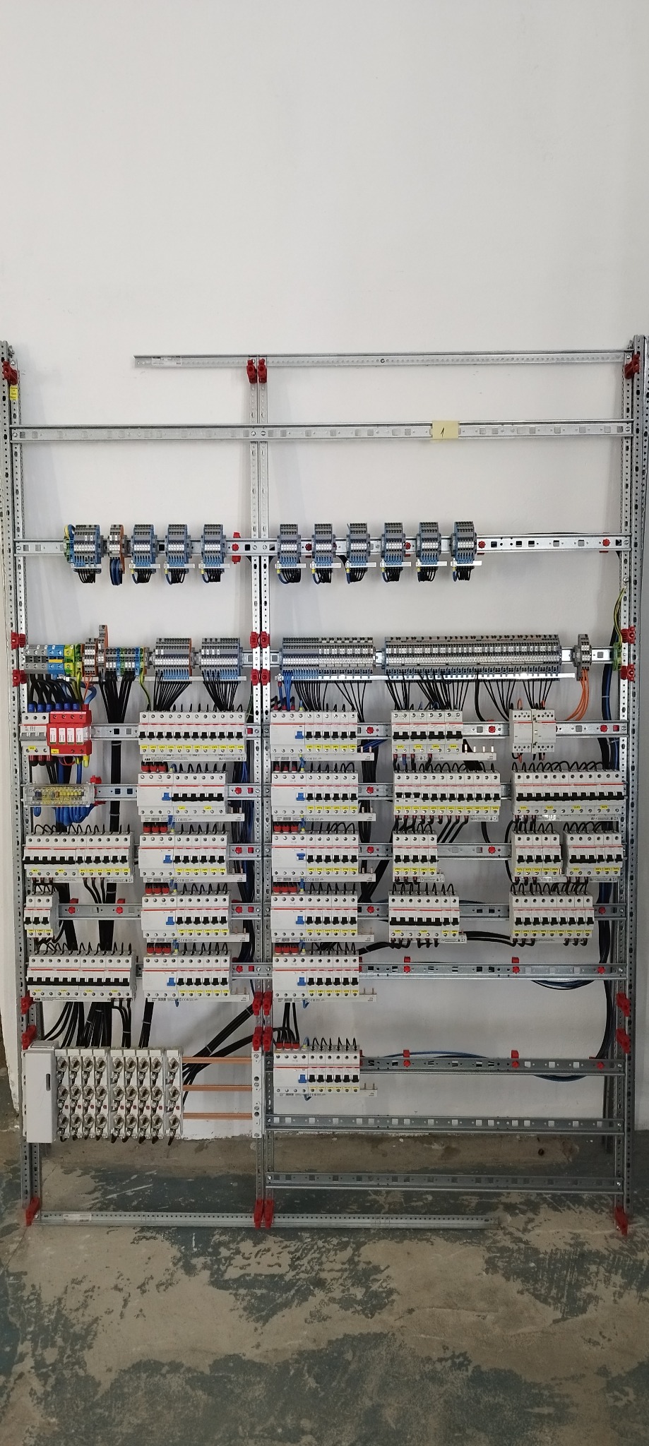 Rozbudowana rozdzielnica elektryczna zamontowana na metalowym stelażu, widoczne liczne zabezpieczenia, złączki i okablowanie, całość na tle białej ściany i surowej podłogi.