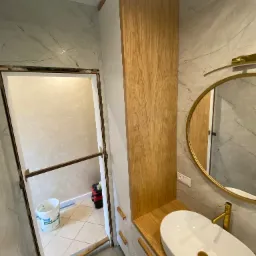 Nowoczesna łazienka w trakcie wykończenia: biała szafka z drewnianymi frontami, owalna umywalka nablatowa z złotą armaturą, okrągłe lustro w złotej ramie na tle marmurowych płytek ściennych. W tle...