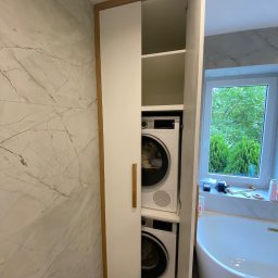 MK Meble Marcin Kustosz - Biała szafa wnękowa z drewna, z ukrytymi pralką i suszarką w łazience z marmurową ścianą. Widoczne okno z zielenią na zewnątrz i fragment wanny.