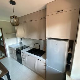 MK Meble Marcin Kustosz - Jasna kuchnia z zabudową meblową, czarnym zlewem i płytą grzewczą, oświetlona lampą w stylu boho, z lodówką Amica wkomponowaną w zabudowę.