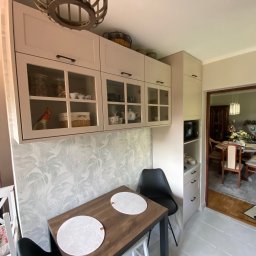 MK Meble Marcin Kustosz - Jasna kuchnia z przeszklonymi szafkami wiszącymi, drewniany stół z okrągłymi podkładkami i czarne krzesła. W tle widoczny salon z jadalnią.