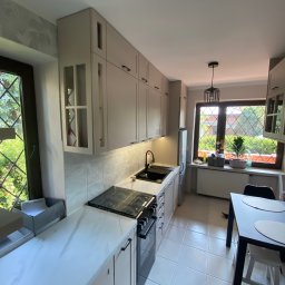 MK Meble Marcin Kustosz - Jasna kuchnia z marmurowym blatem, czarnymi uchwytami i płytą grzewczą, widok na okno z kratką i ogród.