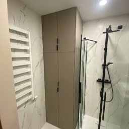 MK Meble Marcin Kustosz - Wysoka, beżowa szafa z czarnymi uchwytami w nowoczesnej łazience z marmurowymi płytkami, obok grzejnika drabinkowego i kabiny prysznicowej z czarnymi elementami.