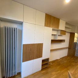 MK Meble Marcin Kustosz - Biało-drewniana zabudowa ściany z szafkami i półkami, dopasowana do grzejnika, z parkietem na podłodze i dekoracyjną ścianą z kamienia w tle.