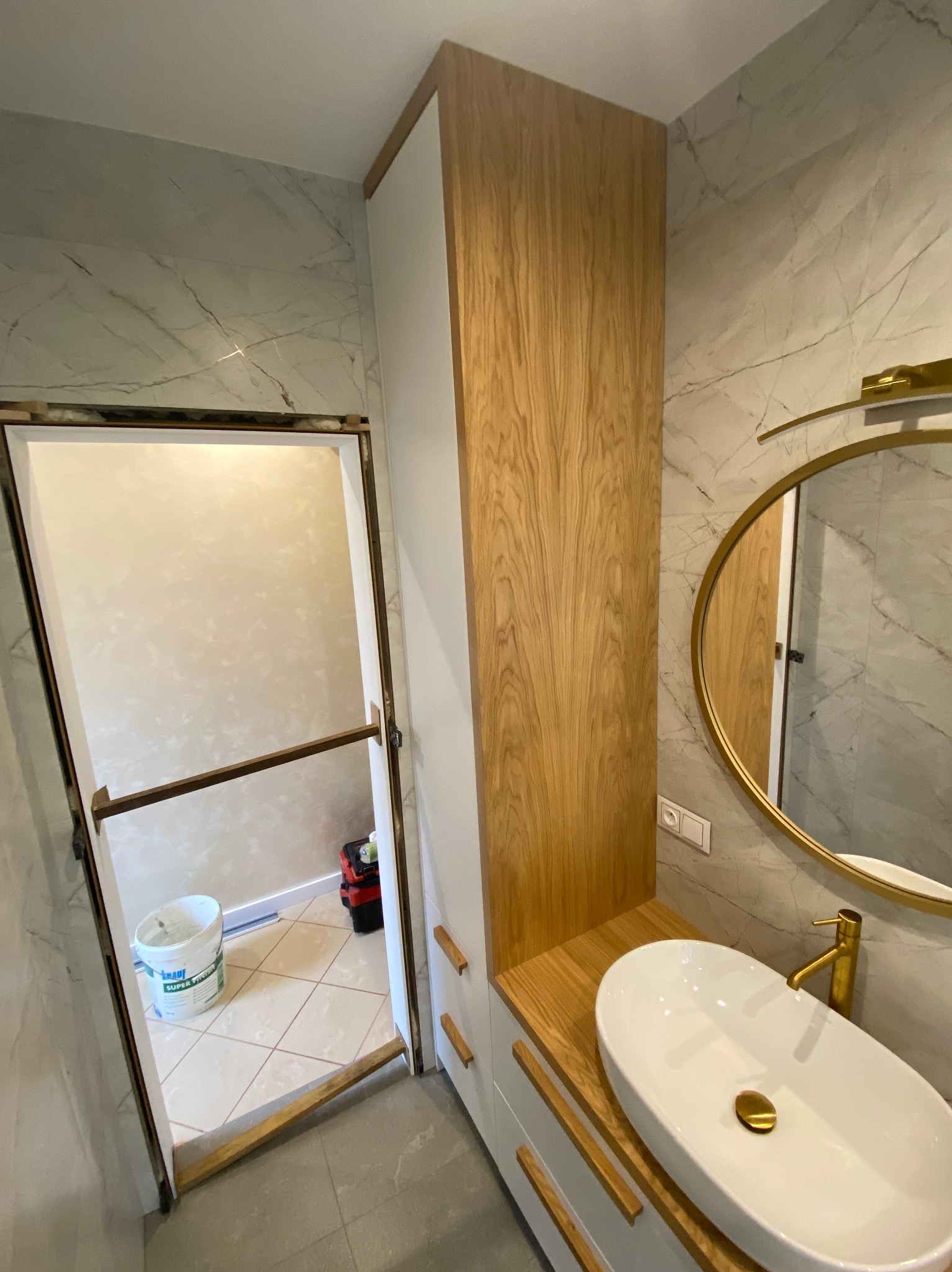 Nowoczesna łazienka w trakcie wykończenia: biała szafka z drewnianymi frontami, owalna umywalka nablatowa z złotą armaturą, okrągłe lustro w złotej ramie na tle marmurowych płytek ściennych. W tle...