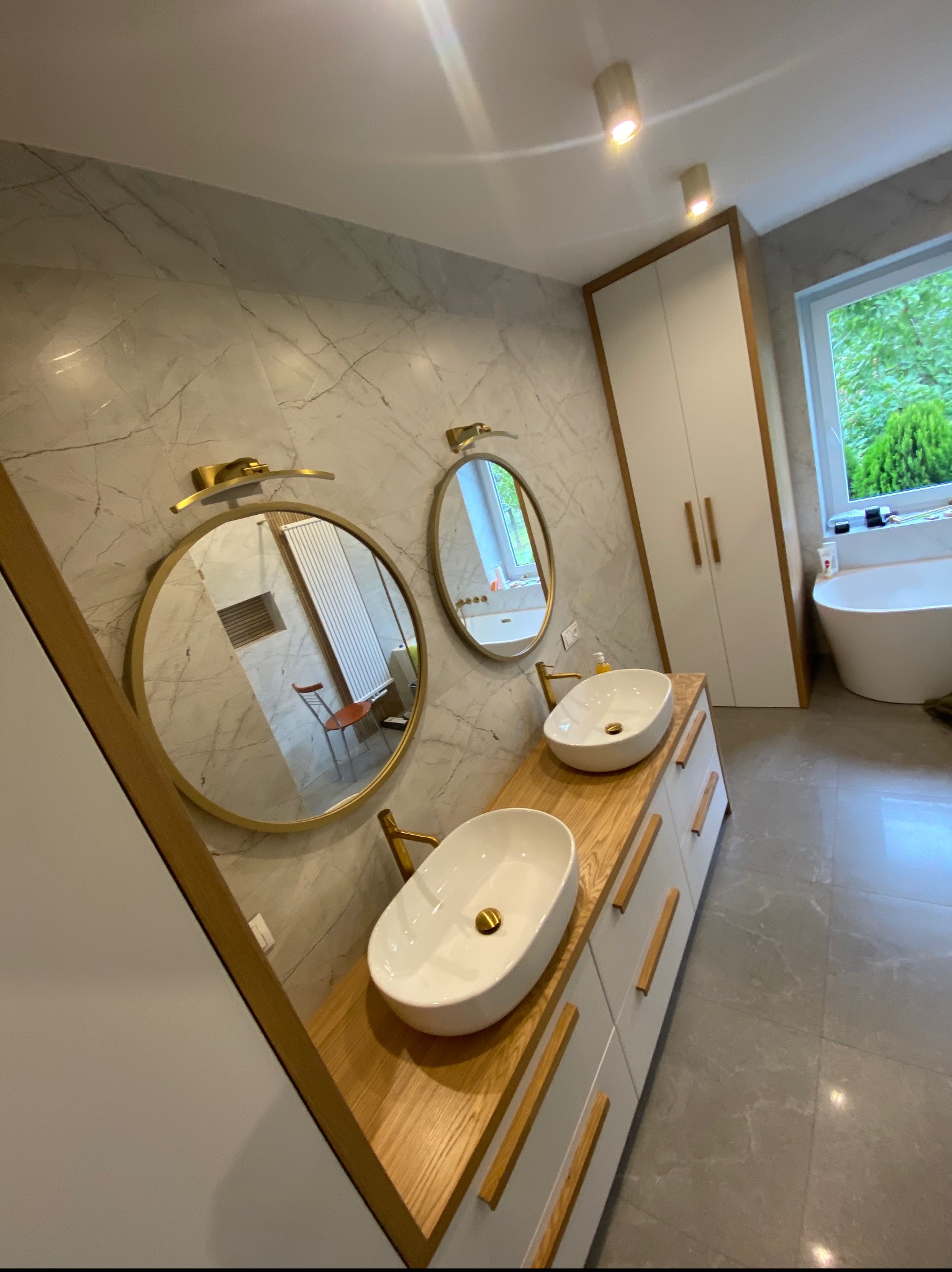 Nowoczesna łazienka z dwoma umywalkami nablatowymi na drewnianym blacie, okrągłe lustra z podświetleniem LED, marmurowe ściany i szafa w zabudowie z drewnianymi uchwytami.