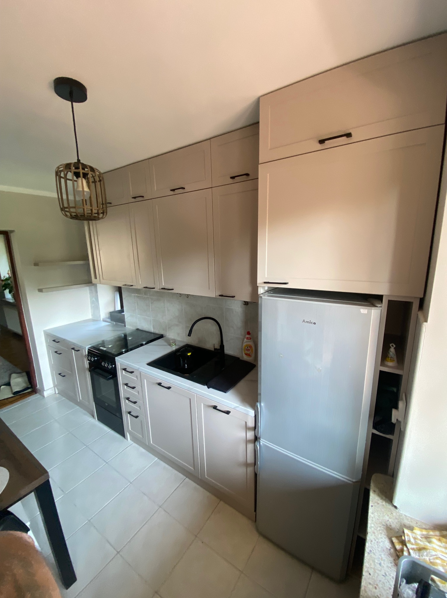 Jasna kuchnia z zabudową meblową, czarnym zlewem i płytą grzewczą, oświetlona lampą w stylu boho, z lodówką Amica wkomponowaną w zabudowę.