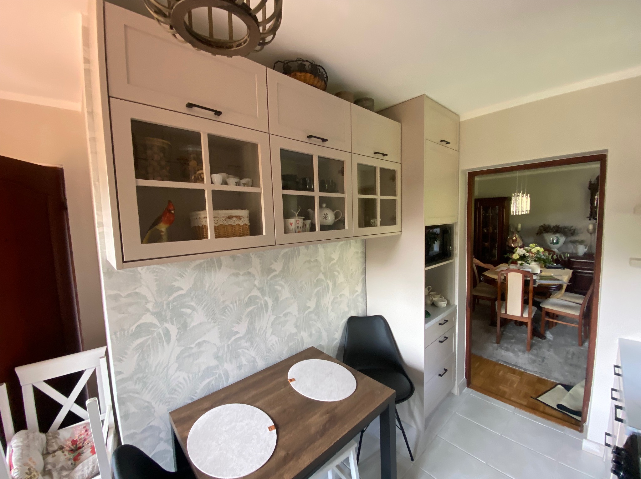 Jasna kuchnia z przeszklonymi szafkami wiszącymi, drewniany stół z okrągłymi podkładkami i czarne krzesła. W tle widoczny salon z jadalnią.