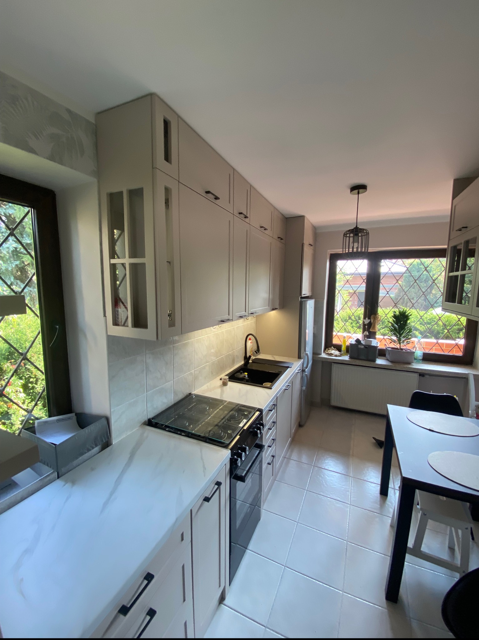 Jasna kuchnia z marmurowym blatem, czarnymi uchwytami i płytą grzewczą, widok na okno z kratką i ogród.