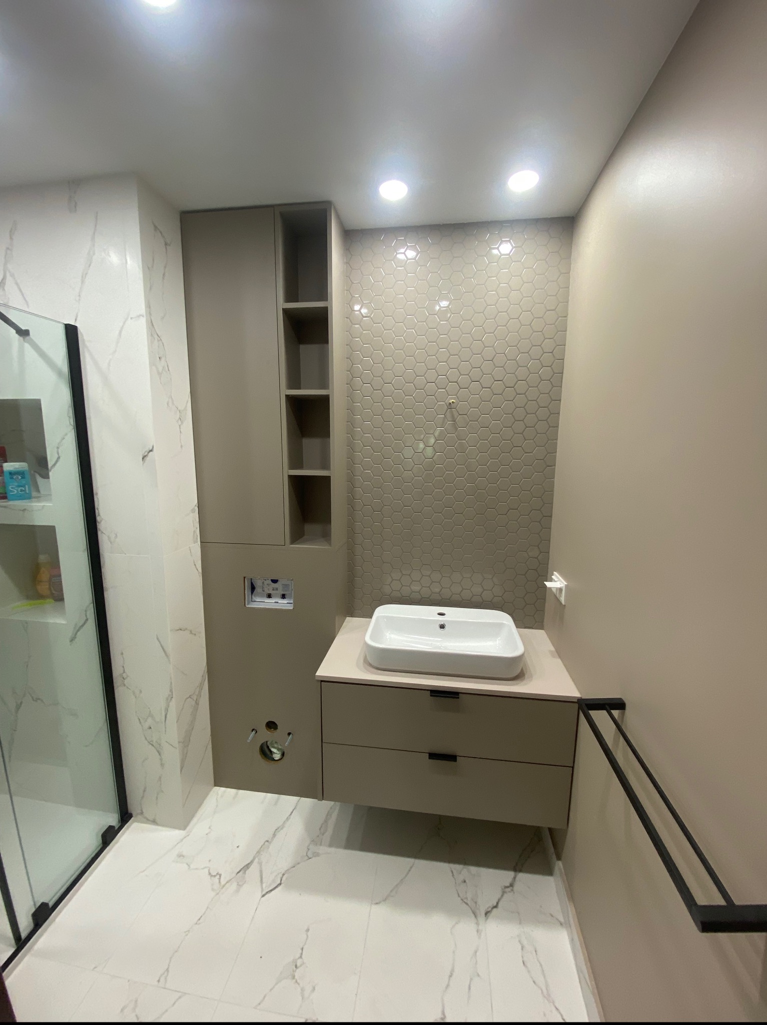 Nowoczesna łazienka z umywalką nablatową, szafką z dwiema szufladami, wysoką szafką z otwartymi półkami i heksagonalnymi płytkami na ścianie. Widoczna kabina prysznicowa z czarnymi ramami...