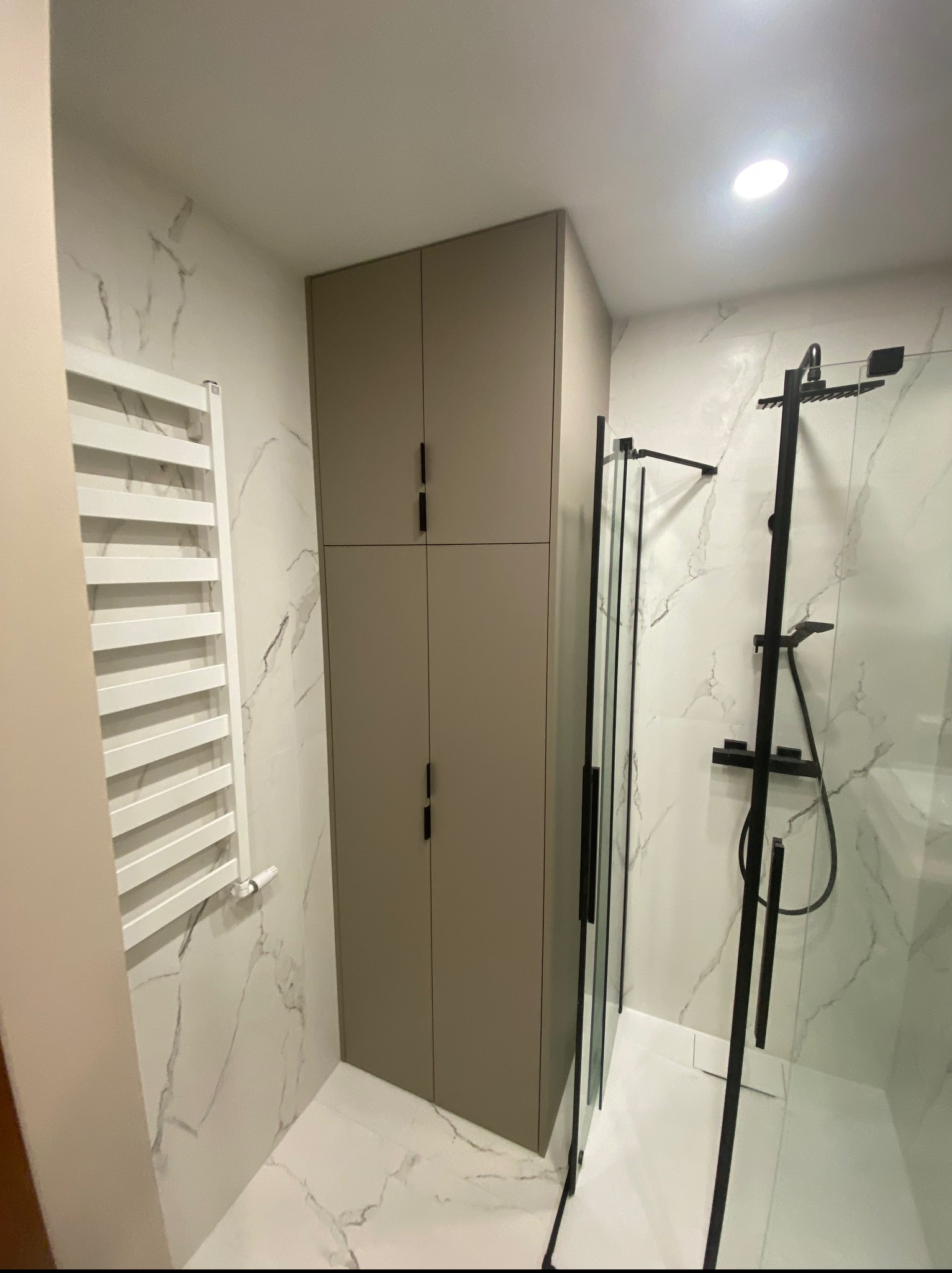 Wysoka, beżowa szafa z czarnymi uchwytami w nowoczesnej łazience z marmurowymi płytkami, obok grzejnika drabinkowego i kabiny prysznicowej z czarnymi elementami.
