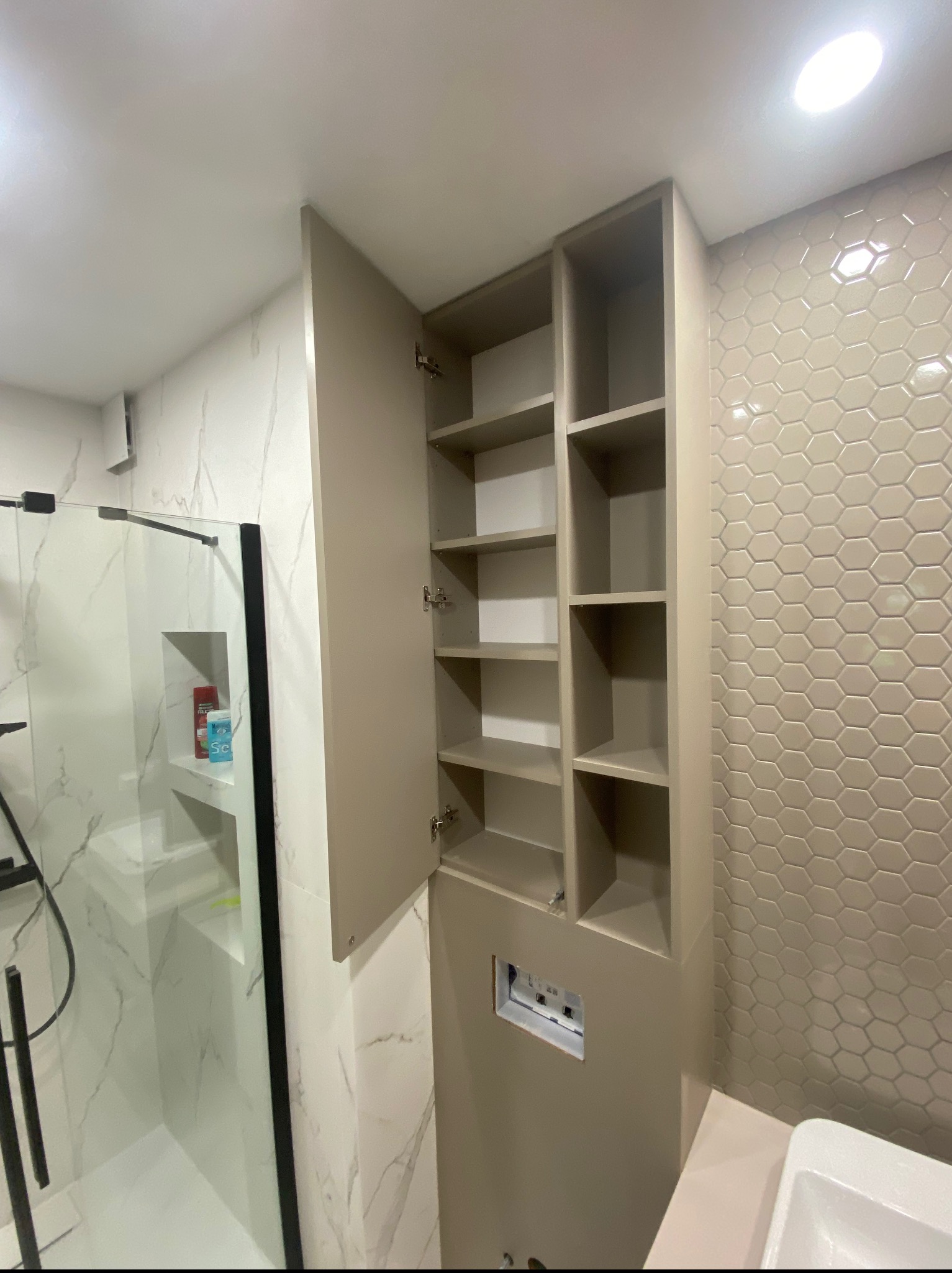 Wysoka szafka łazienkowa na wymiar w odcieniach beżu, z otwartymi półkami i widocznymi zawiasami, zamontowana nad zabudową toalety, na tle heksagonalnych płytek i kabiny prysznicowej z czarnymi...