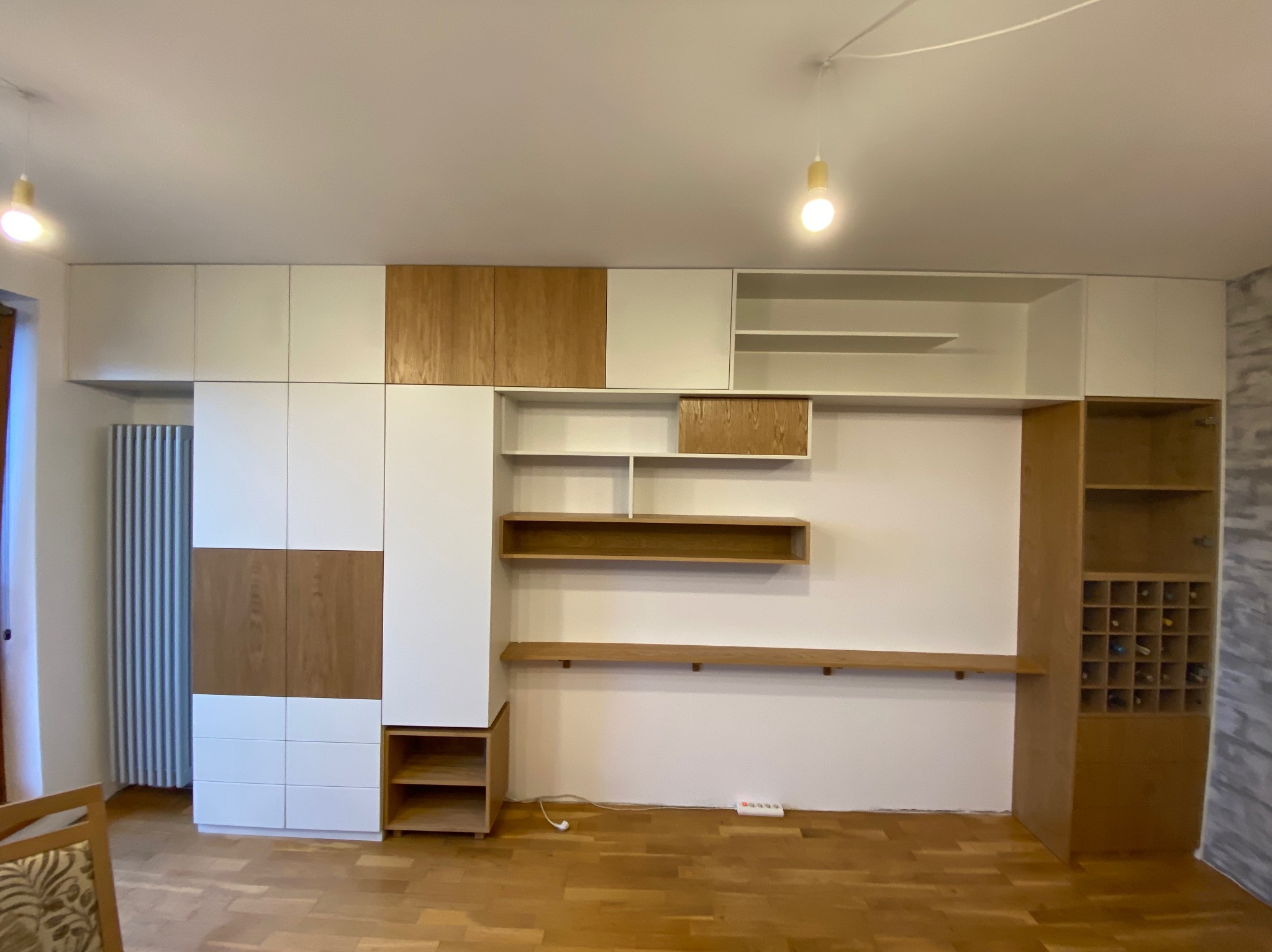 Wielofunkcyjna zabudowa ścienna z białymi i drewnianymi frontami, z półkami, szafkami i regałem na wino, oświetlona wiszącymi lampami.