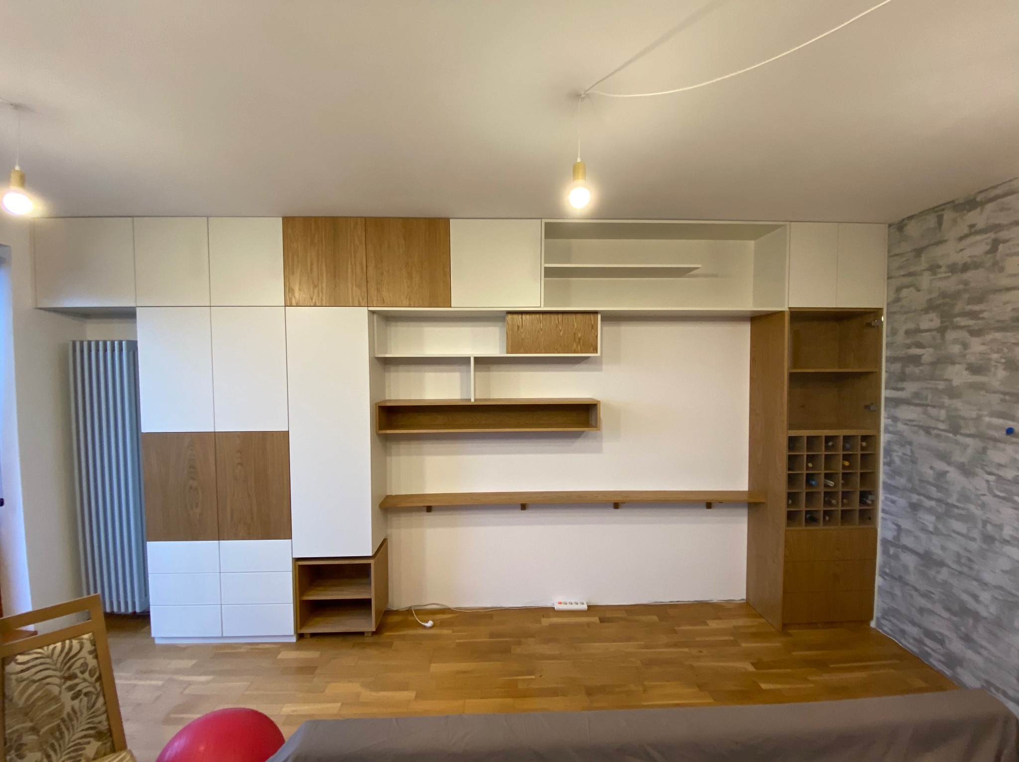 Nowoczesny salon z zabudową meblową: białe i drewniane szafy, półki, regał na wino, oświetlenie punktowe, parkiet, ściana z efektem betonu.