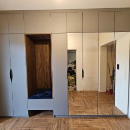 Przemysław Olczyk Meble Olczyk - Szara szafa wnękowa z lustrami i otwartą wnęką z drewnianym wykończeniem, uzupełniona siedziskiem z poduszką, na podłodze ułożonej w jodełkę.