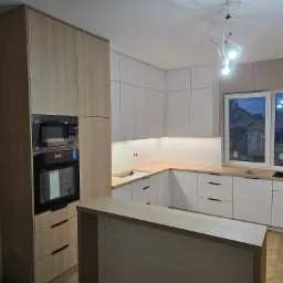 Jasna kuchnia z drewnianymi i białymi szafkami, sprzęt AGD w folii ochronnej, widok na okno z wieczornym krajobrazem. Parkiet na podłodze.