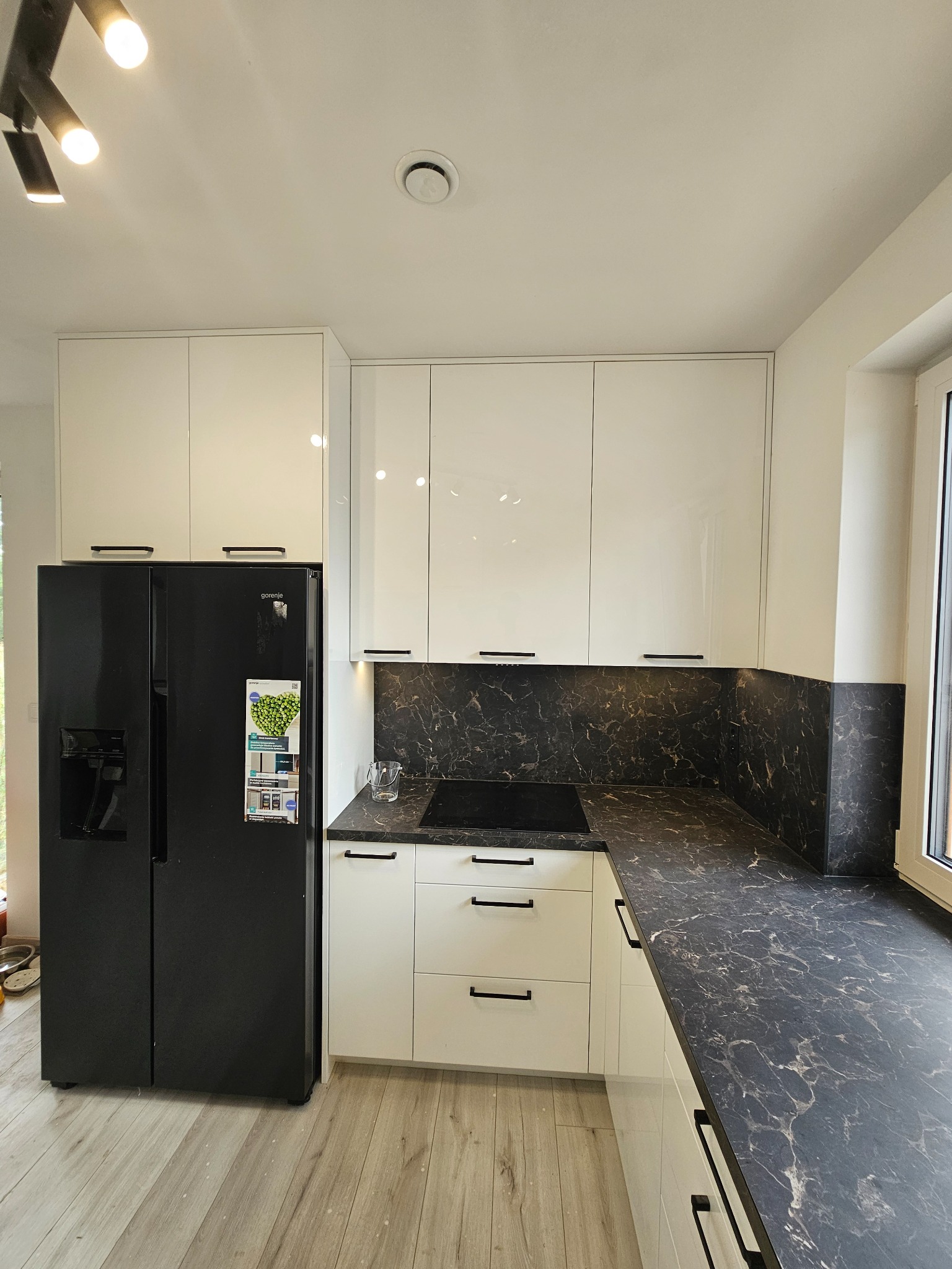 Nowoczesna kuchnia z białymi, lakierowanymi szafkami i czarnym blatem imitującym marmur, czarna lodówka side-by-side, drewniana podłoga.