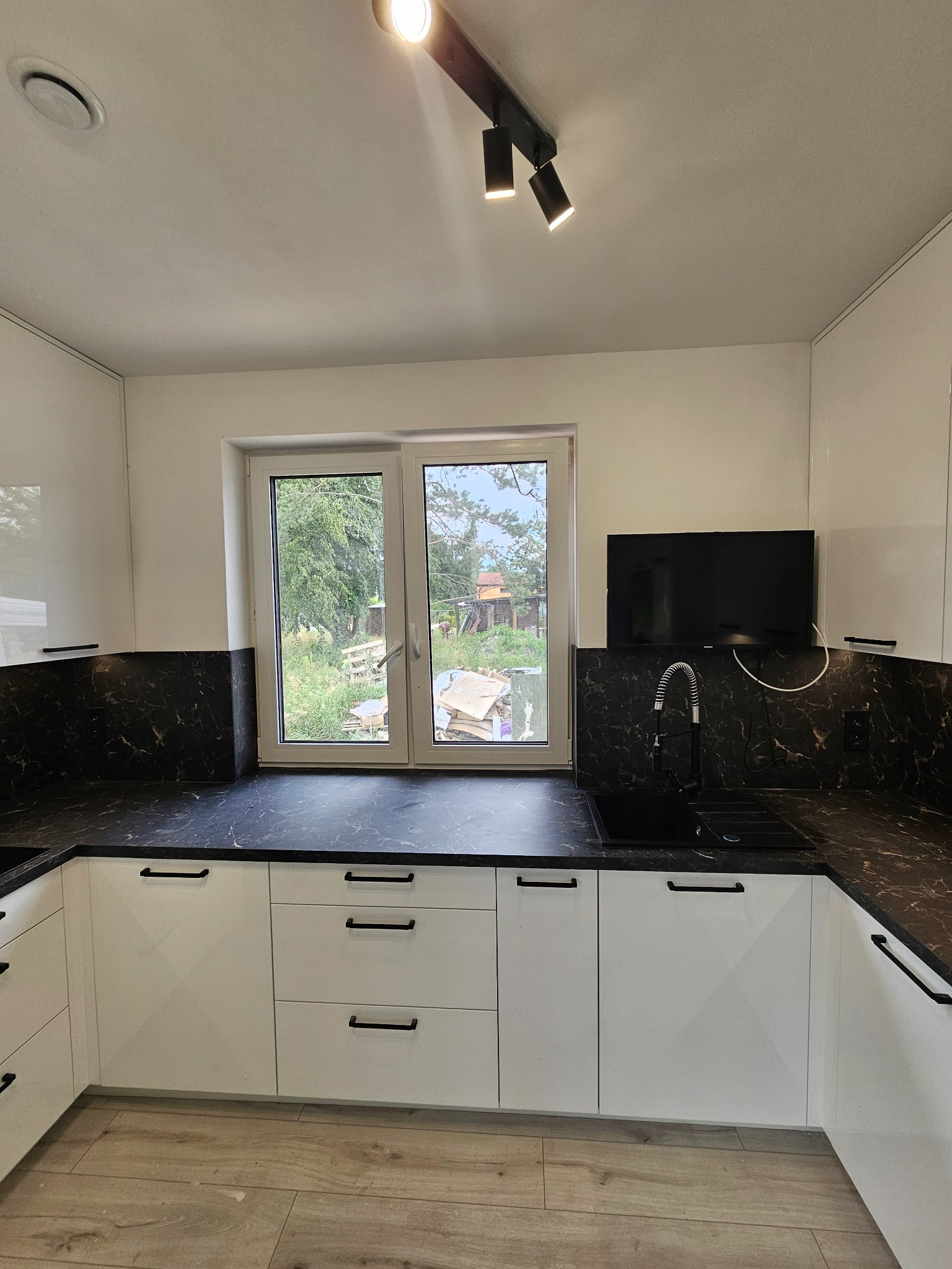 Biała, nowoczesna zabudowa kuchenna z czarnym blatem imitującym marmur, czarnymi uchwytami i oświetleniem punktowym. W tle widoczne okno z widokiem na ogród.