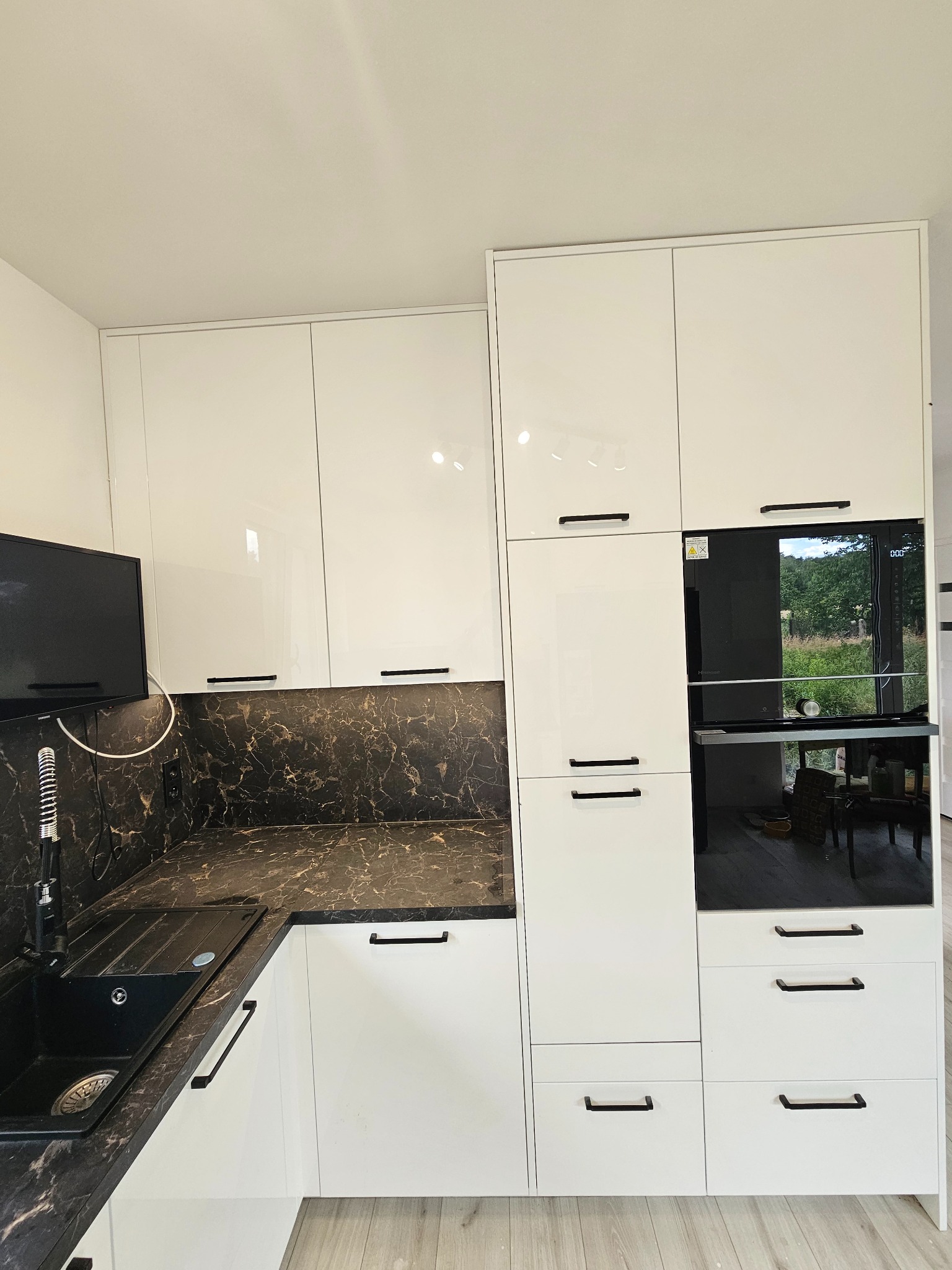 Nowoczesna kuchnia z białymi, lakierowanymi frontami i blatem imitującym czarny marmur w Płoszowie. Uwagę zwraca czarny zlew i bateria oraz zabudowana piekarnik.