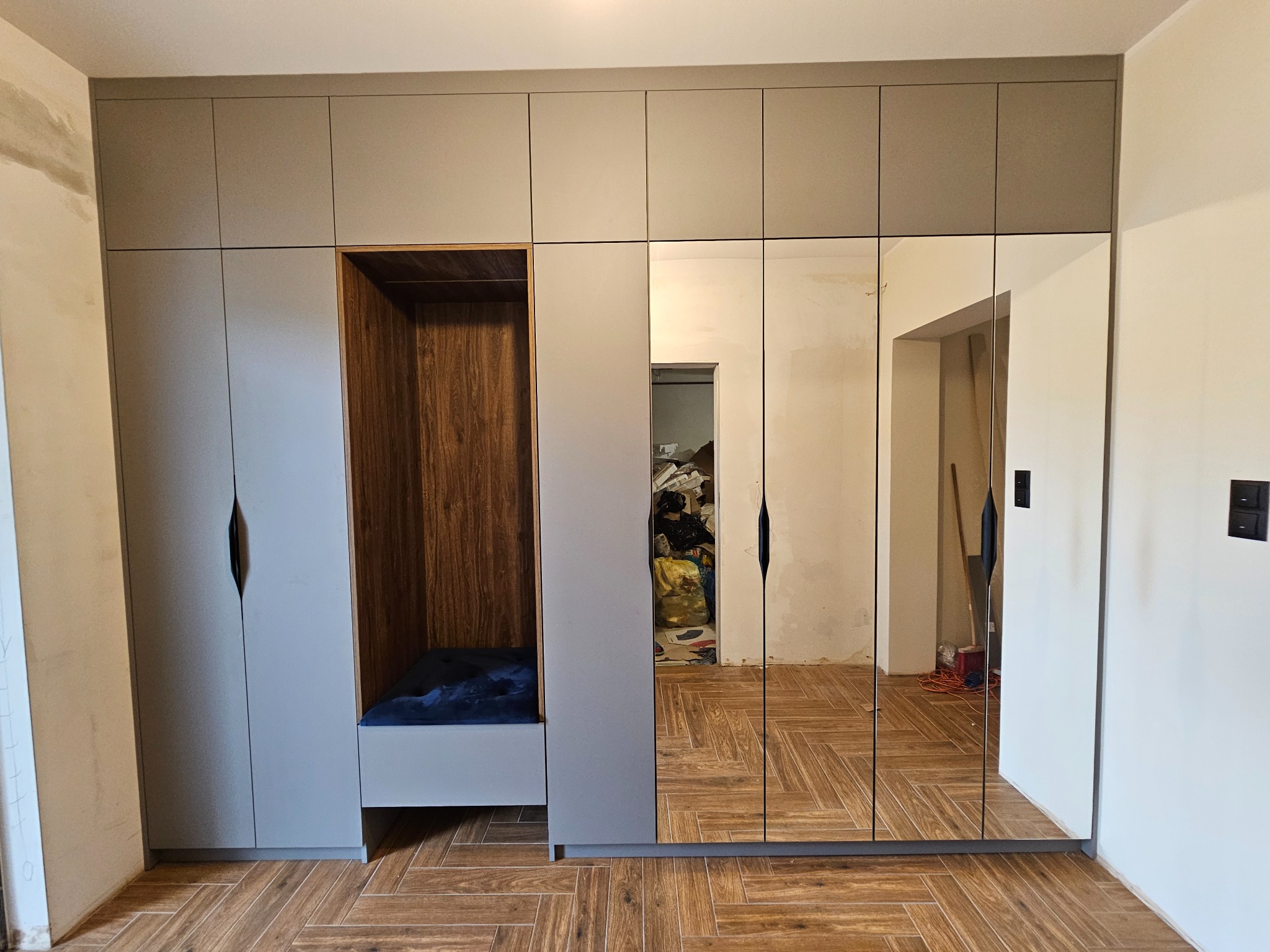 Szara szafa wnękowa z lustrami i otwartą wnęką z drewnianym wykończeniem, uzupełniona siedziskiem z poduszką, na podłodze ułożonej w jodełkę.