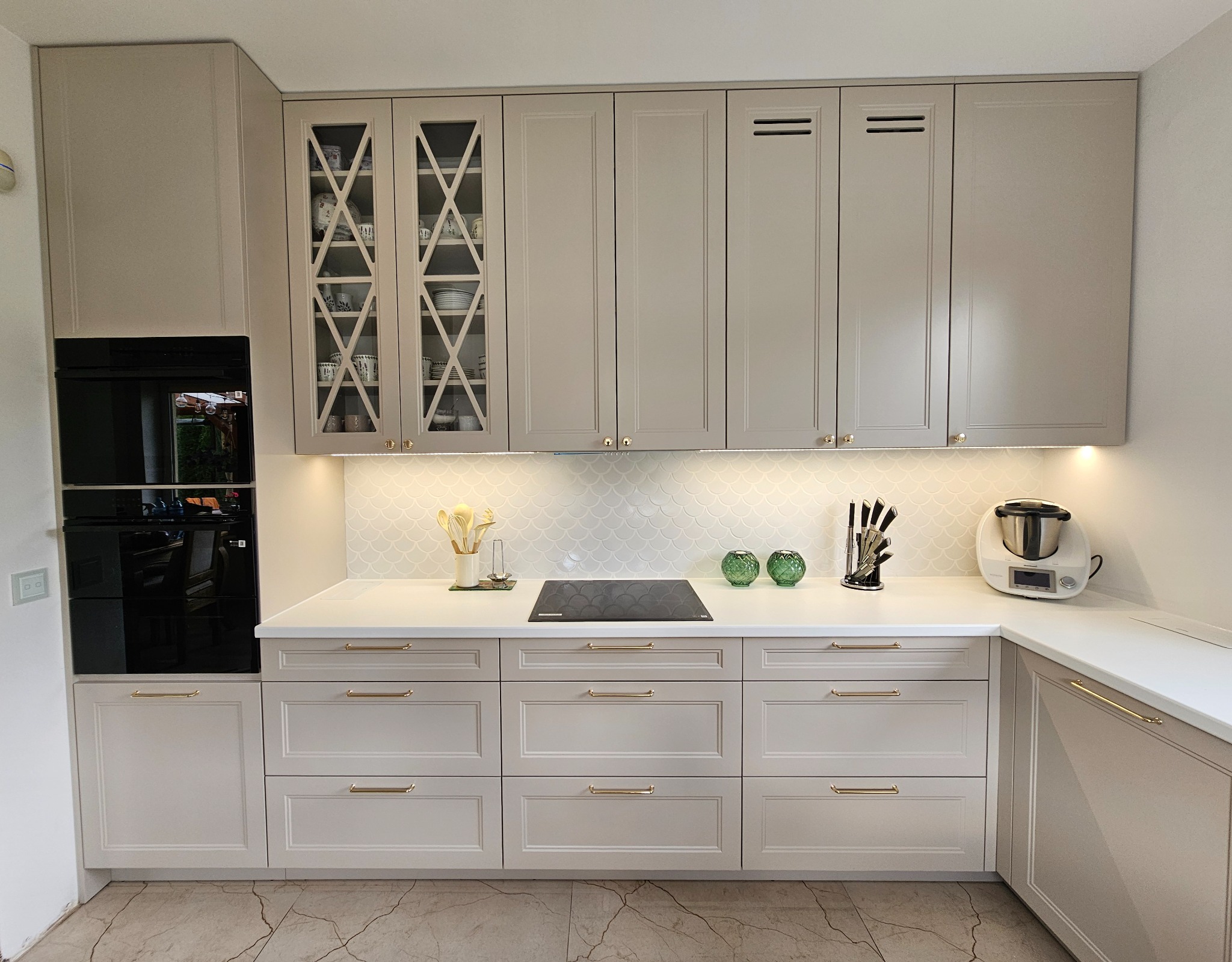 Jasnoszara kuchnia z frezowanymi frontami, złotymi uchwytami i przeszkloną witryną, oświetlona listwą LED pod szafkami wiszącymi, na blacie płyta indukcyjna, robot kuchenny i noże