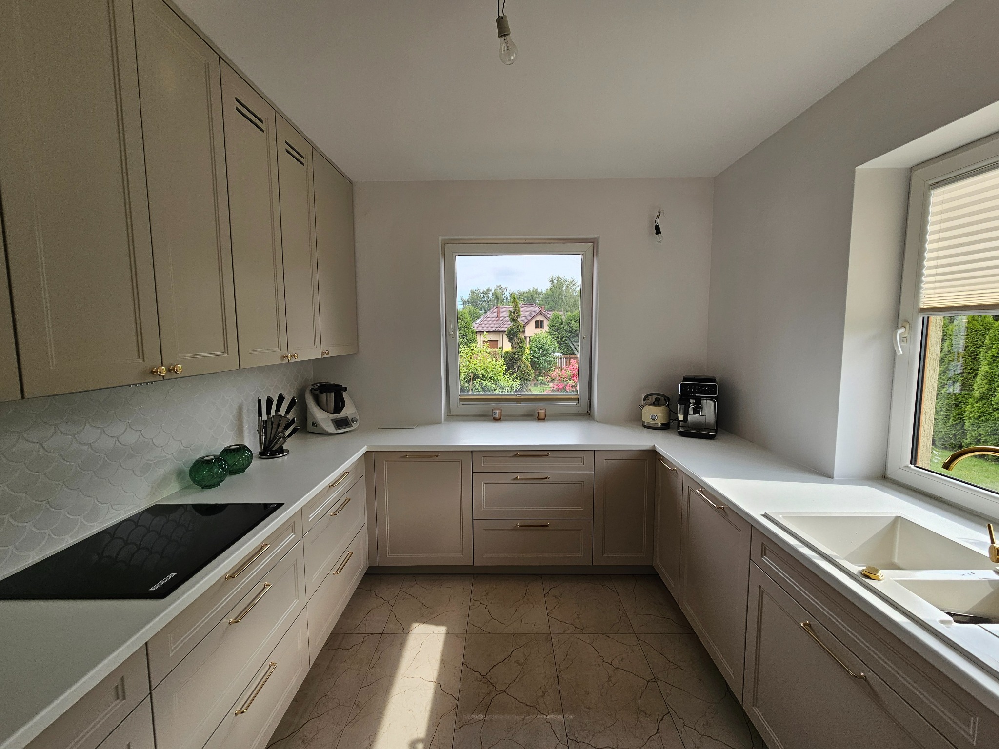Jasna kuchnia w kształcie litery U z beżowymi frontami, złotymi uchwytami i widokiem na ogród przez okno.