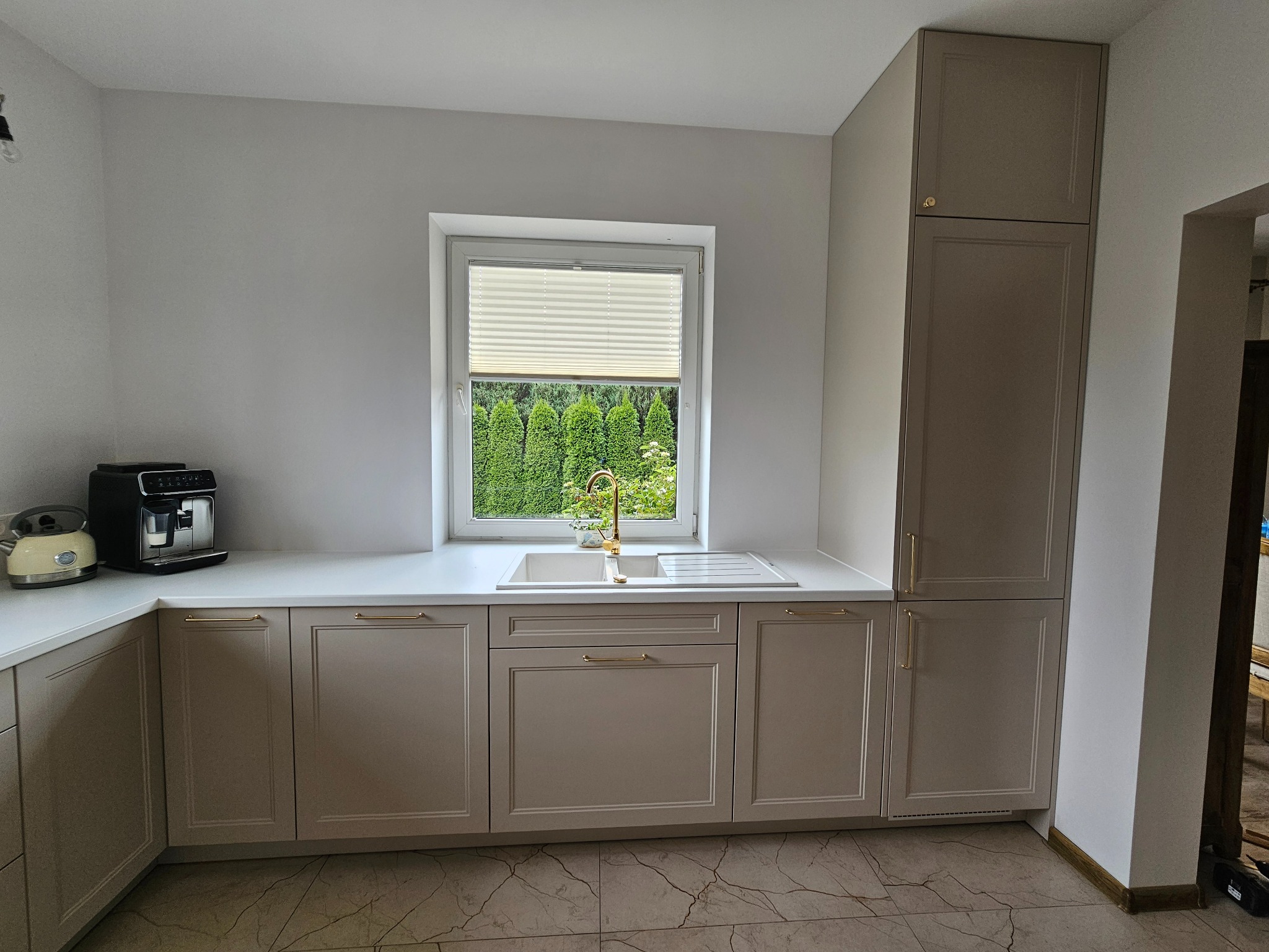 Jasna kuchnia z beżowymi frontami, złotymi uchwytami i armaturą, ekspres do kawy na blacie, widok na zielone tuje za oknem.