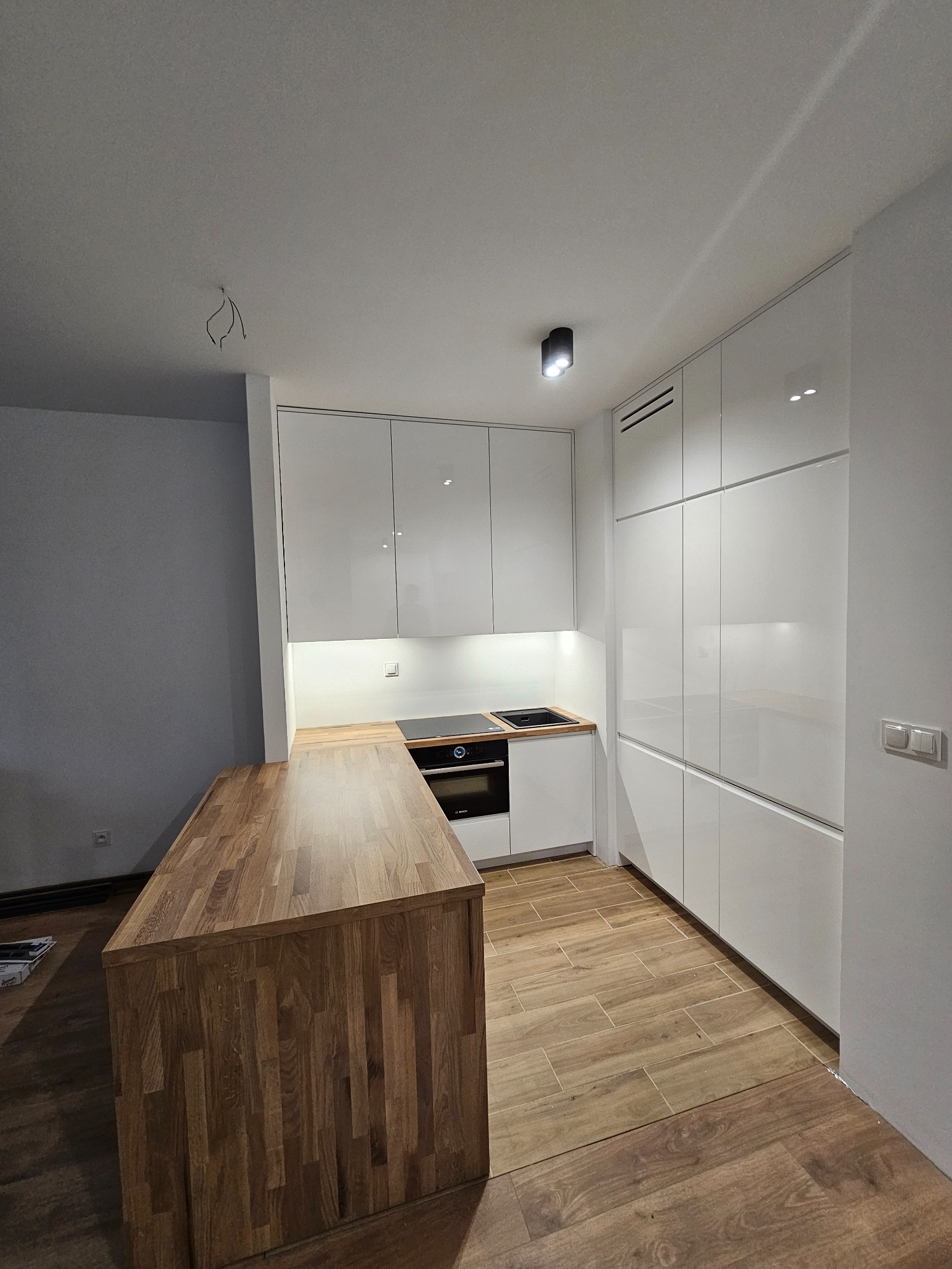 Nowoczesna kuchnia z białymi, lakierowanymi frontami szafek i drewnianym blatem wyspy kuchennej. Widoczne niedokończone podłączenie oświetlenia sufitowego.