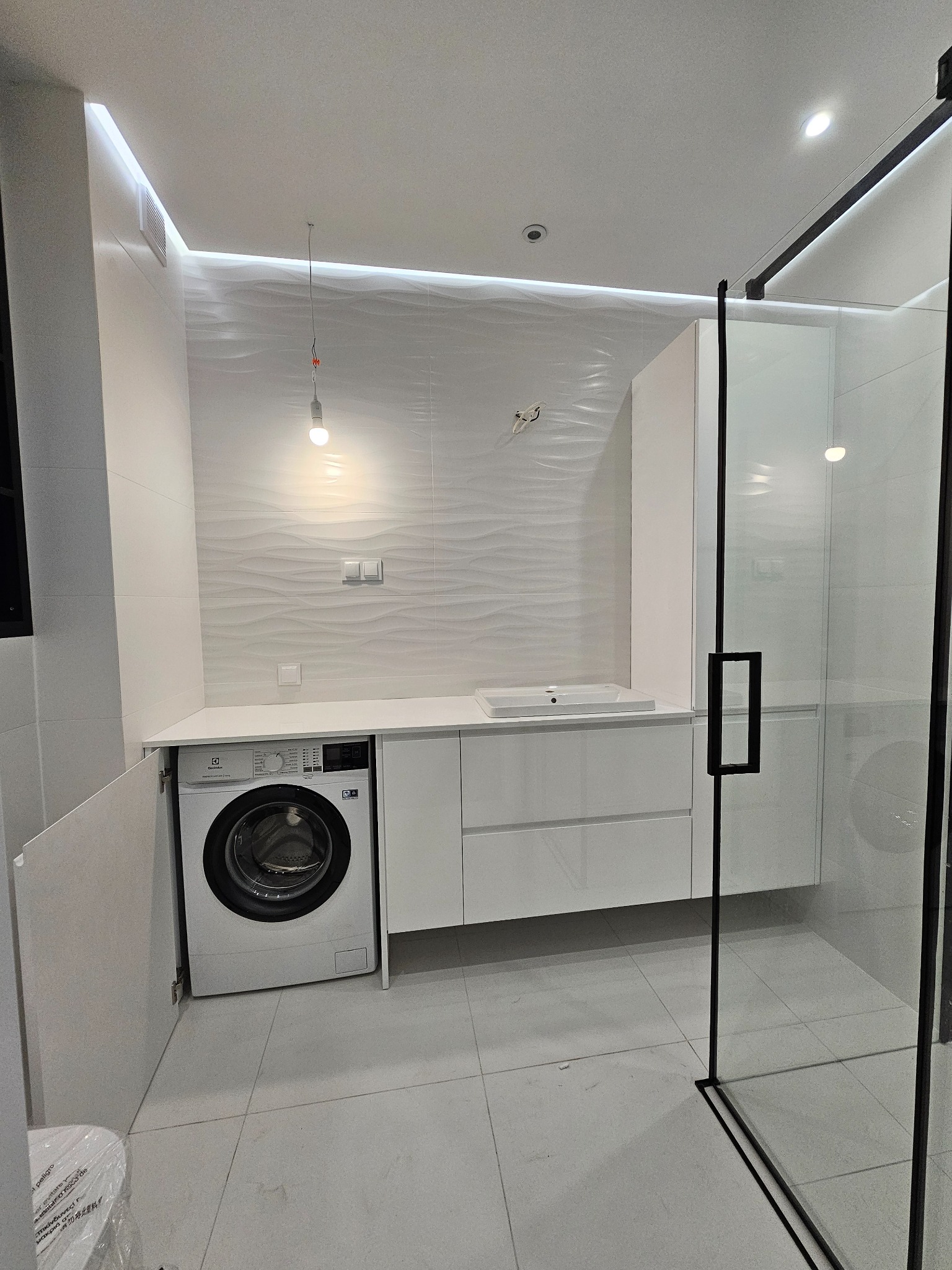 Nowoczesna łazienka z białą zabudową meblową, w której zintegrowano pralkę pod blatem, umywalkę i kabinę prysznicową z czarnymi profilami.