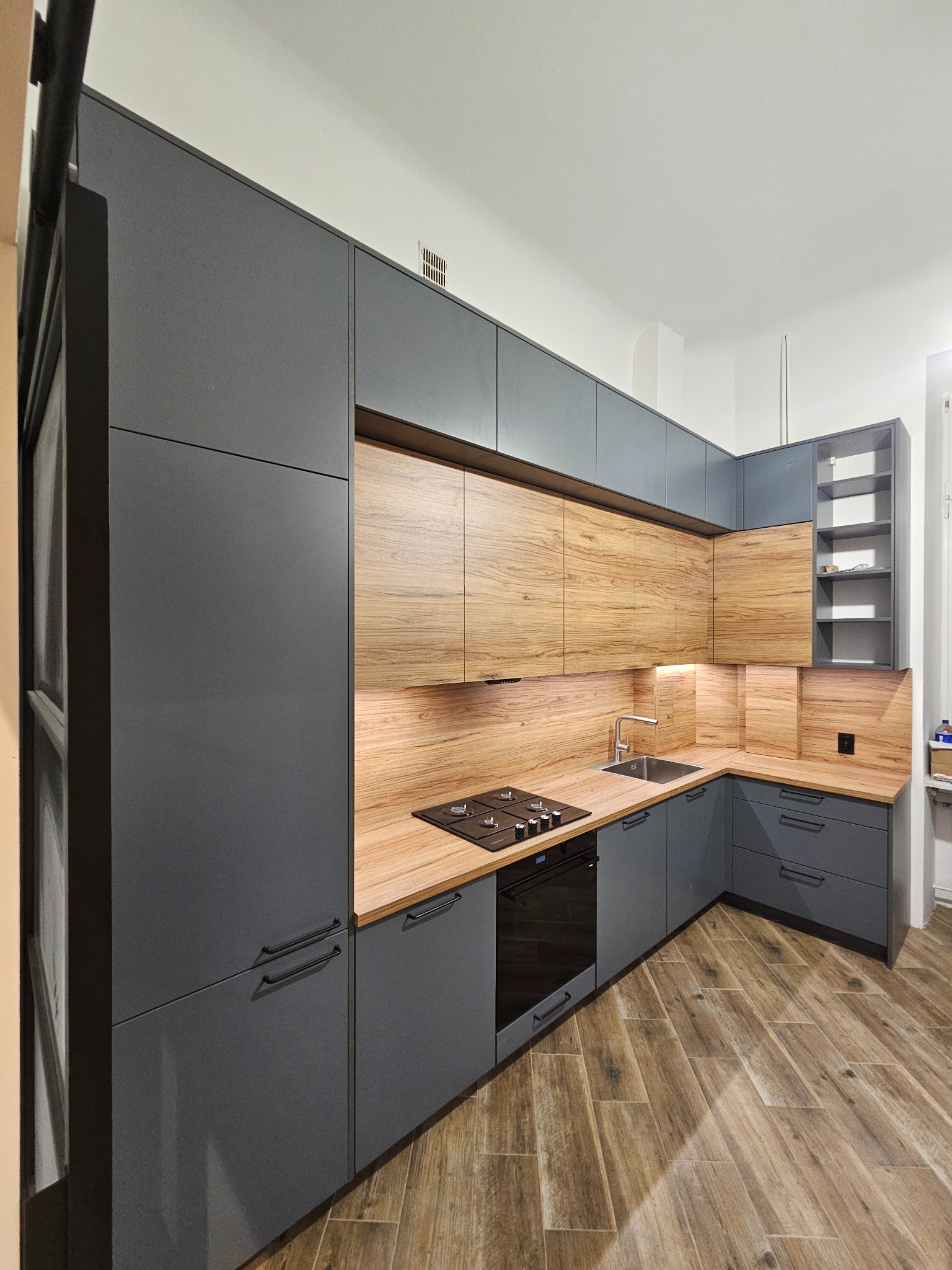 Nowoczesna kuchnia z szarymi szafkami i drewnianymi akcentami, oświetleniem LED pod szafkami wiszącymi i drewnopodobną podłogą.