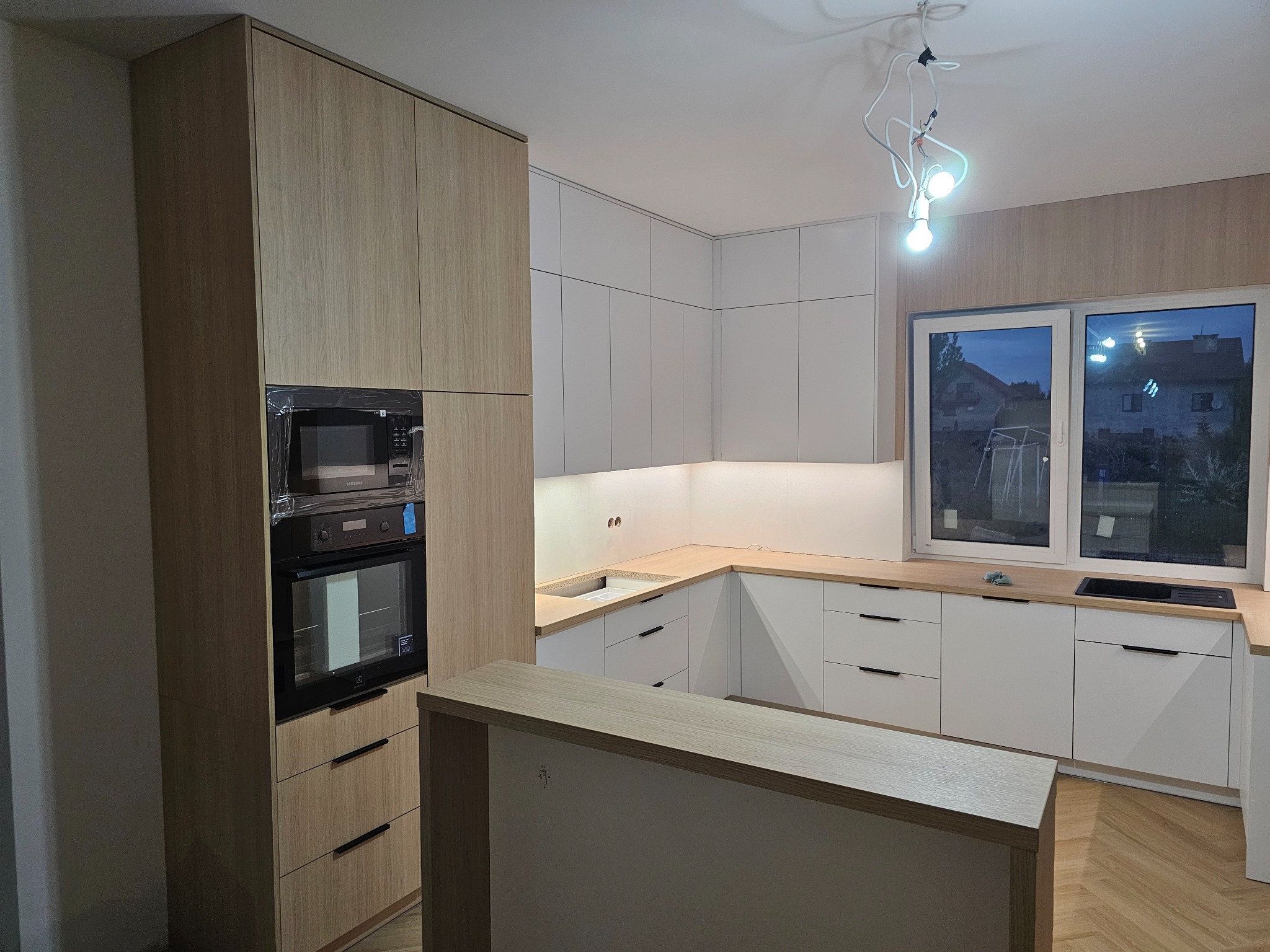 Jasna kuchnia z drewnianymi i białymi szafkami, sprzęt AGD w folii ochronnej, widok na okno z wieczornym krajobrazem. Parkiet na podłodze.