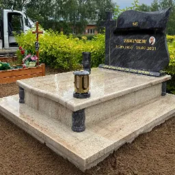 Nowoczesny, dwupoziomowy pomnik z jasnego, polerowanego kamienia z czarną tablicą nagrobną z wygrawerowanym imieniem, nazwiskiem i datami, oraz zniczem na górnej płycie, otoczony świeżo ułożoną...