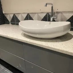 Biała, owalna umywalka nablatowa z chromowaną baterią stojącą na blacie kamiennym z szarymi szafkami pod spodem, wzorzysta ściana w tle.