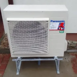 Zewnętrzna jednostka Panasonic na metalowym stelażu, z widoczną naklejką serwisową i oznaczeniem czynnika R32, ustawiona na betonowej powierzchni przy ścianie budynku.