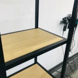 Fragment metalowej konstrukcji regału z drewnianymi półkami w warsztacie, widoczne narzędzia na ścianie w tle.