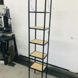 Wysoki, wąski regał metalowy z drewnianymi półkami, wykonany z czarnego metalu, widoczny w warsztacie obok maszyny do obróbki metalu i stosu profili metalowych.