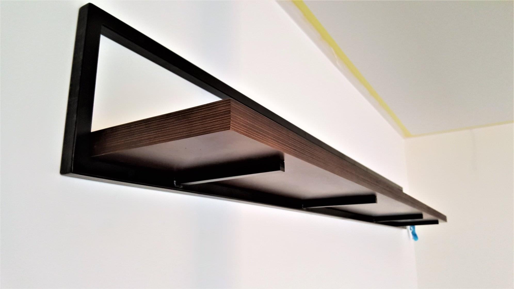 Półka ścienna z ciemnego drewna z metalową ramą, zamontowana na białej ścianie w rogu pokoju, widoczne taśmy malarskie przy suficie.